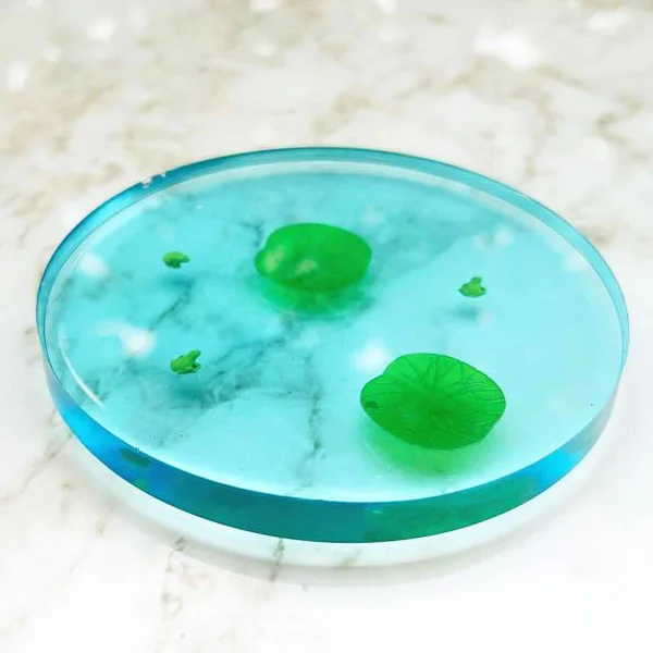 Close up of a blue clear circular coaster with green frogs and lilypads embedded in it